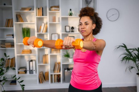 Photo for Healthy young woman doing exercises with dumbbells at home. Fit young female doing weights workout indoors in living room. - Royalty Free Image