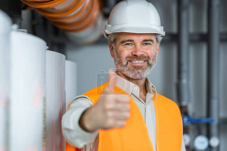 Photo for Smiling handsome caucasian worker in protective working clothes and with helmet on head near boiler and giving thumb up while standing in factory. - Royalty Free Image