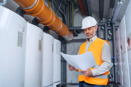 Photo for Dedicated factory worker standing next to boiler and holding drafts and papers. Worker is dressed in protective uniform, having hardhat. old man with blueprints working in a factory - Royalty Free Image
