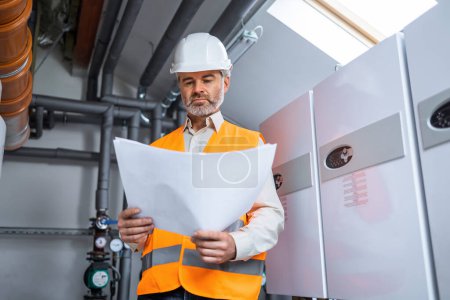Photo for Portrait of maintenance mature senior gray haired engineer worker doing quick inspection of production in heating plant. old man with blueprints working in a factory - Royalty Free Image