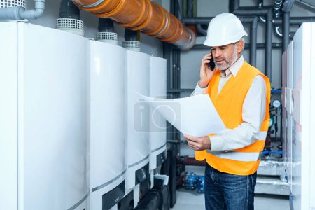 Photo for The senior gray haired technician checking the heating system in the boiler room with paper draft in hand - Royalty Free Image