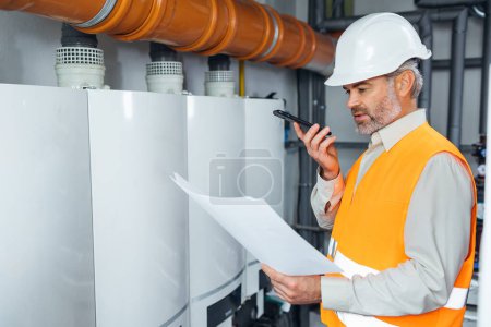 Photo for Handsome engineer with blueprints on industrial plant. Man with technical drawings in hands on oil and gas factory using phone for audio message - Royalty Free Image