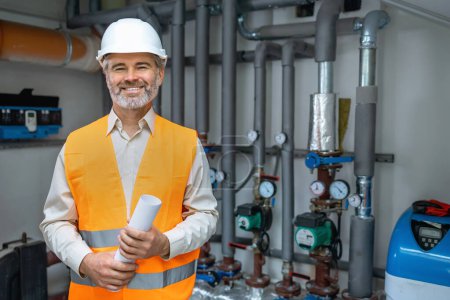 Photo for Senior gray haired Dedicated smiling factory worker standing next to pipes with gas pressure. Worker is dressed in protective uniform, having hardhat - Royalty Free Image