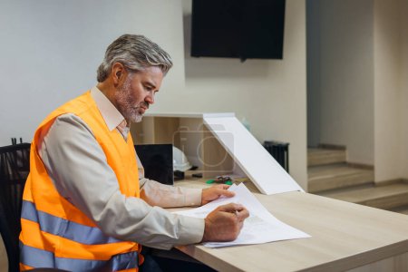 Photo for Builder, engineer in reflective vest uniform on desk in office looking through papers or sketches. - Royalty Free Image