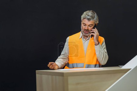 Photo for Competent architect male sitting on desk indoors talking on phone. bearded mature man wearing uniform orange reflective vest - Royalty Free Image