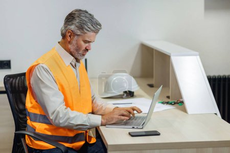 Photo for Gray haired attractive construction bearded man in vest working with laptop, work on Home building project - Royalty Free Image