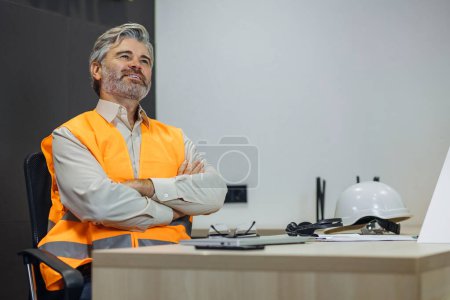 Photo for Front view of pensive adult male engineer wearing orange reflective vest thinking sit in office - Royalty Free Image