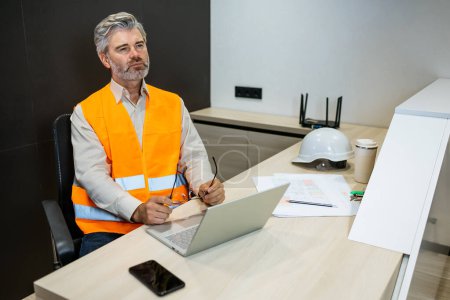 Photo for Pensive frowning construction engineer sitting at office desk with blueprints and laptop looking away - Royalty Free Image
