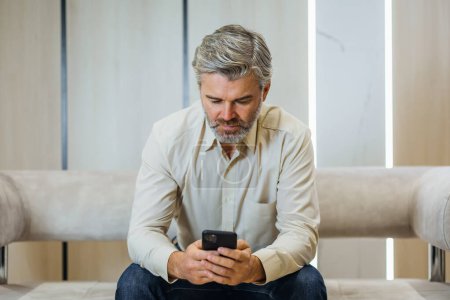 Téléchargez les photos : Homme âgé en vêtements formels en utilisant le téléphone assis sur le canapé au bureau moderne de luxe - en image libre de droit