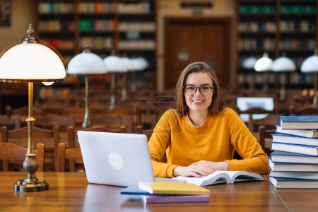 Foto de Hermosa joven estudiante sonriente en ropa juvenil en el portátil en la biblioteca con libros cerca - Imagen libre de derechos