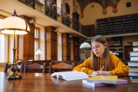 Foto de Chica sentada en una silla en la mesa, hace sus deberes en la biblioteca, escribe palabras de un libro en un cuaderno. Educación, conocimiento, librería - Imagen libre de derechos