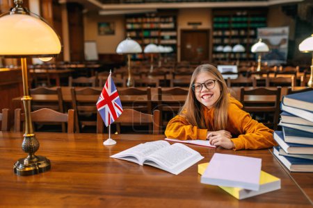 Foto de Una colegiala enfocada en un suéter amarillo se sienta en una mesa en la biblioteca de la escuela con gafas en el telón de fondo de la bandera británica. - Imagen libre de derechos