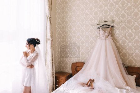 Téléchargez les photos : Une mariée coûteuse se tient dans une robe de chambre près de la fenêtre et regarde le marié. matin, préparation pour les vacances. - en image libre de droit