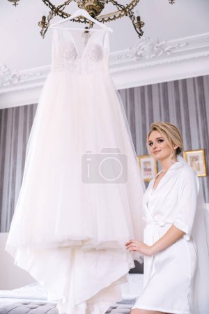Téléchargez les photos : Une mariée rayonnante se tient près d'une robe de mariée suspendue au plafond. - en image libre de droit