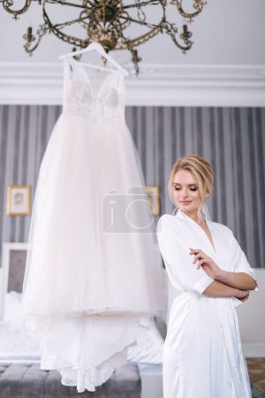 Téléchargez les photos : Préparation matinale de la mariée. fille dans une belle robe près d'une robe de mariée suspendue sur un cintre à l'intérieur. - en image libre de droit