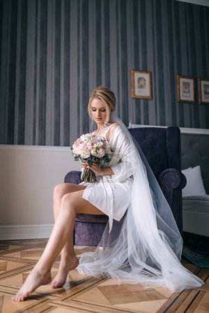 Téléchargez les photos : La mariée tient un bouquet de mariage entre ses mains. Assis dans une chaise en robe blanche et voile. - en image libre de droit