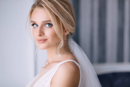 Foto de Hermosa novia con cabello rubio en vestido de novia elegante posando en la habitación en la mañana de boda - Imagen libre de derechos