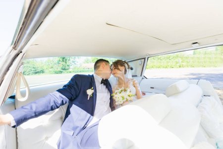 Photo for The groom in a blue suit and the bride in a beautiful dress are sitting in the back seat of a car and kissing. - Royalty Free Image