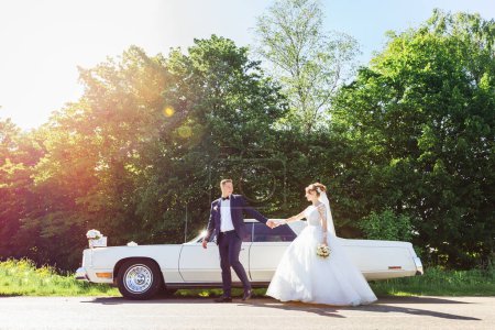 Téléchargez les photos : Le marié tient la main de la mariée et la regarde. jeunes mariés près de voiture rétro. - en image libre de droit