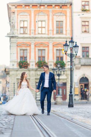 Foto de La novia y el novio se miran y se toman de la mano en la plaza de la ciudad. casco antiguo con vías de tranvía y farolas. - Imagen libre de derechos