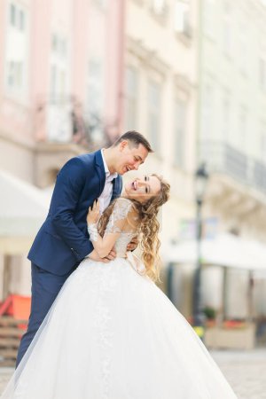 Photo for The groom hugs the bride by the waist and they smile on the city street. - Royalty Free Image