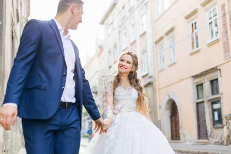 Foto de El novio con traje azul toma la mano de la novia y camina por las calles de la ciudad. linda novia sonriendo. - Imagen libre de derechos