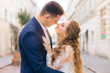 Téléchargez les photos : Les jeunes mariés fermèrent les yeux et s'embrassèrent dans la rue de la ville. la lumière du soleil entre les mariés. - en image libre de droit