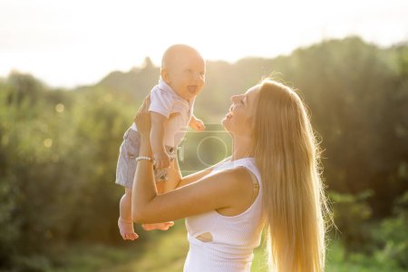 Photo for Happy young mother playing and having fun with her little baby son on sunshine warm spring or summer day. Beautiful sunset light in the apple garden or in the park. - Royalty Free Image