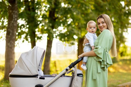 Téléchargez les photos : Jeune mère marchant avec son bébé sur les mains et poussette au parc le jour ensoleillé - en image libre de droit