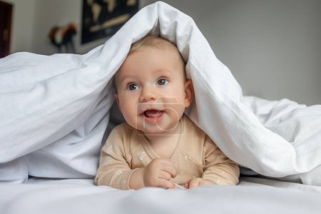 Téléchargez les photos : Magnifique bébé joue sur le lit dans un drap léger, se cachant sous une couverture blanche, jouer avec maman cacher et chercher - en image libre de droit