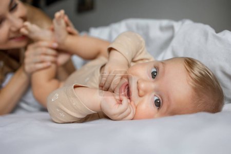 Photo for Portrait of happy baby playing on bed look at camera, mother on background kissing legs. selective focus - Royalty Free Image