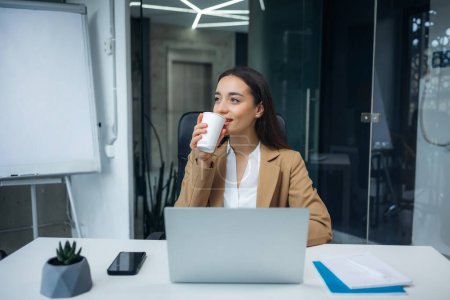 Photo for Young business woman wearing beige suit drinking coffee in office. Happy smiling female professional working get ready for productive day - Royalty Free Image