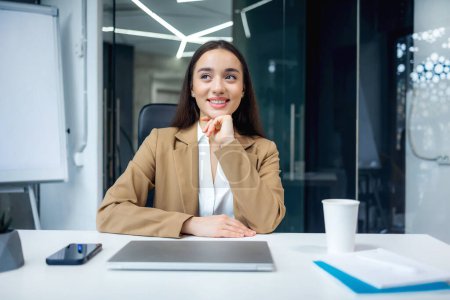 Attractive businesswoman looking aside while sitting at work desk with laptop in office. Confident pretty caucasian lady dreaming of opportunities indoors