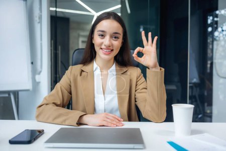Téléchargez les photos : Femme d'affaires heureuse montrant signe geste correct assis au bureau et en utilisant un ordinateur portable sur fond de bureau. recommander un travail en entreprise ou un cours éducatif webinaire - en image libre de droit
