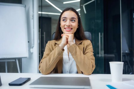 Photo for Young businesswoman reliving happy memories during a coffee break sitting at her desk in the office looking to the side with a smile of contentment and pleasure - Royalty Free Image