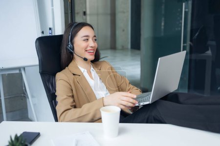 Foto de Mujer alegre sentada en el lugar de trabajo con piernas en el escritorio y divirtiéndose mientras tiene llamada en auriculares inalámbricos. - Imagen libre de derechos