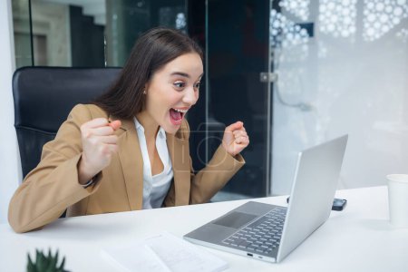 Excited cheerful young woman using laptop computer at office getting good news, feeling joy