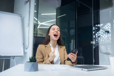 Overjoyed woman screaming and holding phone, celebrating success or online lottery win, job promotion, excited businesswoman showing yes gesture, laughing, sitting at work desk