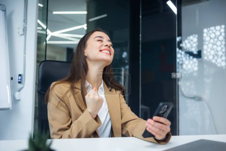 Excited woman sit at desk make win gesture holding phone, online lottery, happy businesswoman overjoyed get mail at phone being promoted at work, amazed read good news