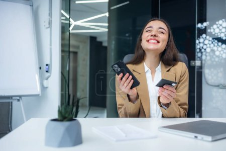 Young happy employee business woman in shirt sit work at office desk with pc laptop using mobile cell phone hold credit bank card do online shopping order