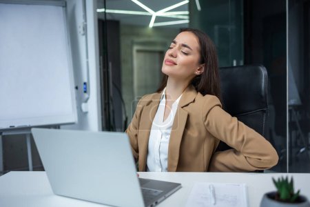 Téléchargez les photos : Femme d'affaires caucasienne travaillant avec un ordinateur portable dans le cabinet se sentant mal au dos sévère tout en étant assis sur une chaise de bureau à l'intérieur. - en image libre de droit
