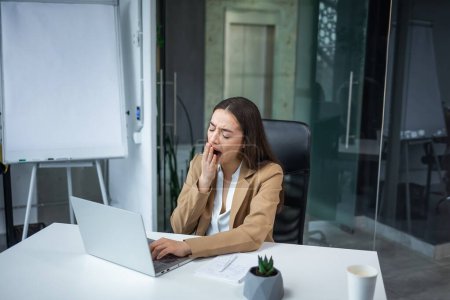 Téléchargez les photos : Fatigué somnolent femme caucasienne bâiller tout en travaillant au bureau pendant le travail avec ordinateur portable. Femme d'affaires surmenée vêtue d'un élégant costume beige assis sur le lieu de travail - en image libre de droit