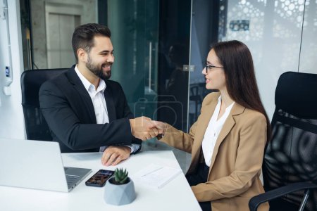 Photo for Caucasian man and woman in formal wear making hands shaking after serious conversation at modern office. Two partners sitting together at desk and having successful deal about new business project. - Royalty Free Image