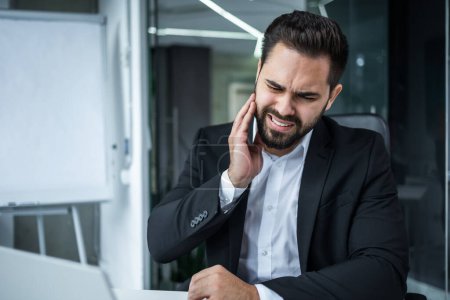 Photo for Toothache in the workplace. A young man, a businessman, an employee, has a teeth pain at work. He is sitting at a desk in the office, holding his cheek - Royalty Free Image