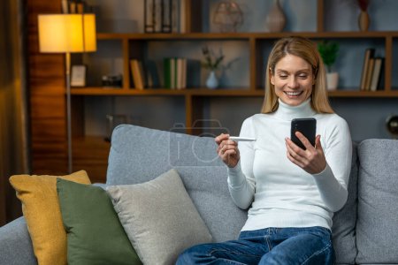 Téléchargez les photos : Smiling and excited adult woman checking her recent pregnancy test, sitting on gray couch at home, copy space - en image libre de droit