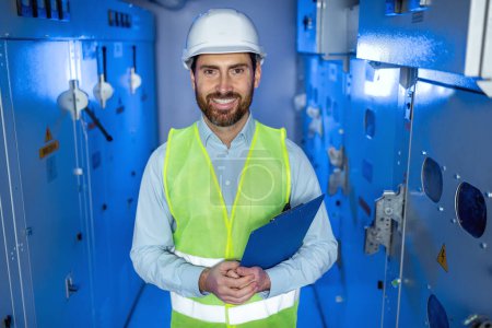 Positive factory worker in white hard hat and green safety vest hold clipboard. Electrician man looking at camera with happy face at electric technical blue room.