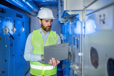 Technology engineer in vest and safety helmet holding laptop and entering data in an electrical switchboard technical room