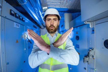 Portrait of male electrical engineer technician worker in white hard hat substation worker gestures to halt against a backdrop of blue light, high voltage, and malfunctioning electrical equipment.