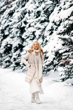 Foto de Moda joven mujer rubia sonriente en invierno. De pie entre los árboles nevados en el bosque de invierno. Abrigo beige, bufanda blanca en estilo boho - Imagen libre de derechos