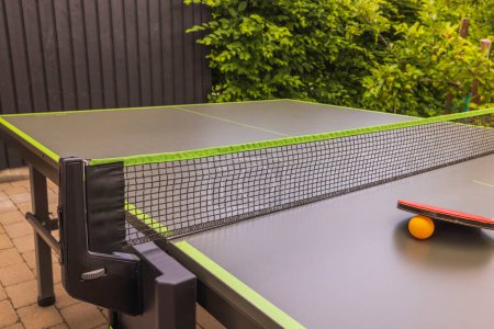 Photo for View of ping-pong racket and orange plastic ball on green netted tennis table in courtyard of villa. Concept of active lifestyle. Sweden. - Royalty Free Image